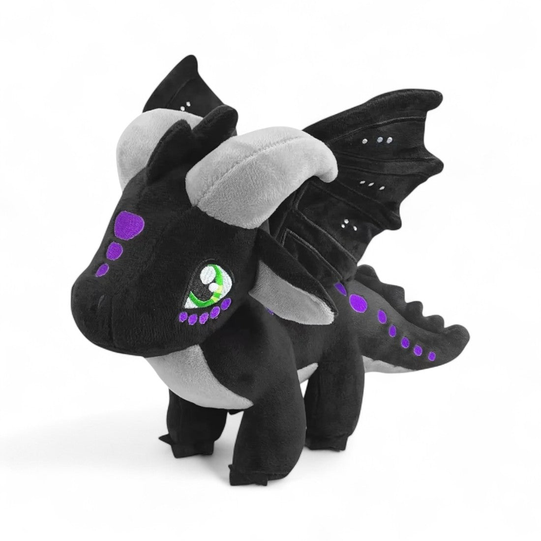 Peluche dragon noir aux détails violets, grandes cornes grises, ailes noires décorées de points lumineux, yeux verts expressifs, sur fond blanc