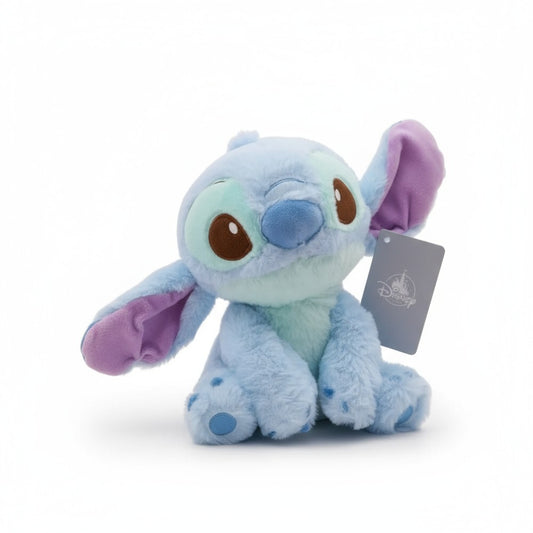 Peluche Stitch bleu tendre aux grandes oreilles et au pelage pastel, assis, sur fond blanc.