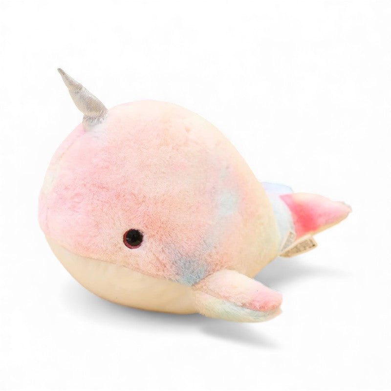 Peluche narval ronde et douce, aux couleurs pastel rose, bleu et jaune, avec une petite corne argentée sur la tête et deux nageoires de chaque côté, posée sur un fond blanc.