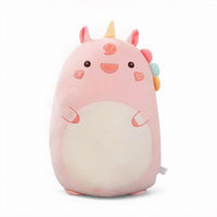 Grand coussin peluche licorne rose, avec corne jaune, crinière multicolore, joues en forme de cœur et grand ventre crème, posé sur un lit dans une chambre lumineuse.