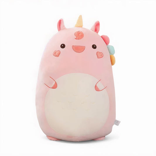 Grand coussin peluche licorne rose, avec corne jaune, crinière multicolore, joues en forme de cœur et grand ventre crème, posé sur un lit dans une chambre lumineuse.