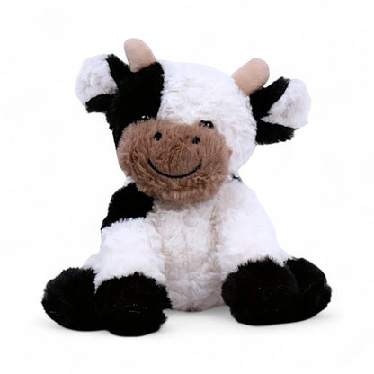 Vache en peluche blanche et noire, museau beige souriant, cornes courtes, oreilles rondes, assise sur fond blanc.