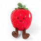 Peluche représentant une fraise rouge avec des jambes marrons et des feuilles vertes sur le dessus, avec deux yeux et un petit sourire brodés style kawaii, assise sur un fond blanc.