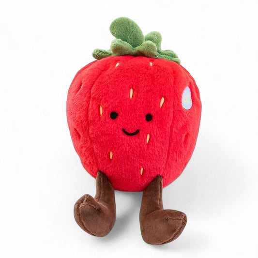 Peluche représentant une fraise rouge avec des jambes marrons et des feuilles vertes sur le dessus, avec deux yeux et un petit sourire brodés style kawaii, assise sur un fond blanc.
