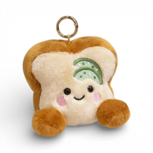 Un petit porte-clé en peluche représentant une tranche de pain toast beige avec une garniture concombre verte sur le dessus, visage souriant aux grands yeux ronds et joues roses, petites pattes marron des deux côtés, anneau métallique pour attache, sur fond blanc.