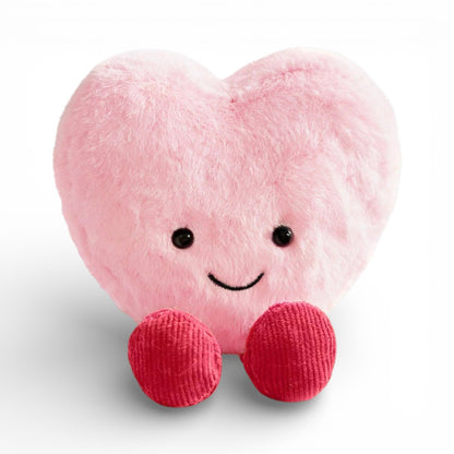 Une peluche en forme de cœur rose doux et duveteux, avec un grand sourire joyeux, deux yeux ronds noirs brillants et deux petites pattes en tissu tricoté rouge en bas, posée sur un fond blanc uni