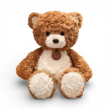 Un ours en peluche brun clair duveteux au ventre blanc doux, avec yeux ronds noirs expressifs, museau adorable, petites oreilles rondes, pattes potelées et médaillon cuir rond autour du cou, posé sur fond blanc uni.